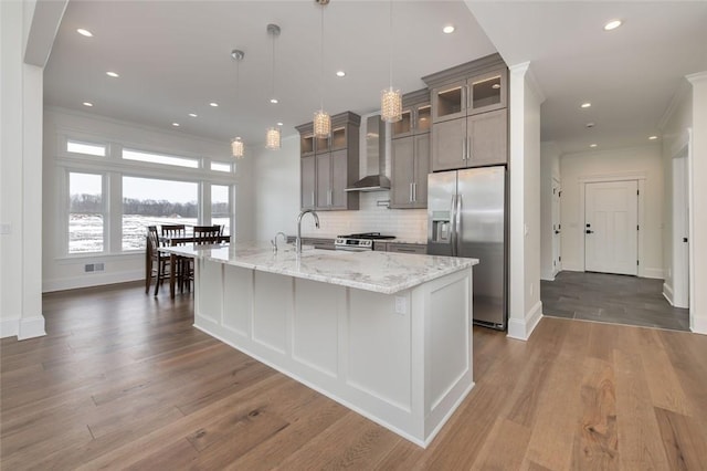 kitchen featuring a center island with sink, stainless steel appliances, backsplash, ornamental molding, and wall chimney exhaust hood