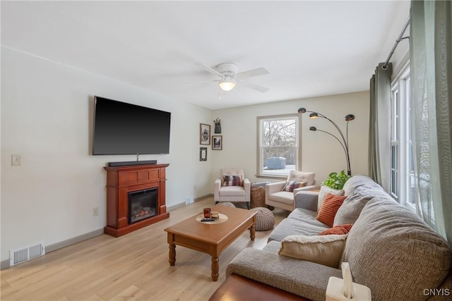 living room with visible vents, light wood-style floors, a glass covered fireplace, ceiling fan, and baseboards