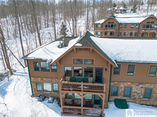 snow covered house featuring a balcony, stone siding, and stucco siding