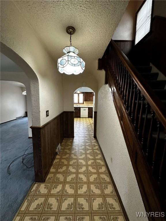 corridor featuring arched walkways, a wainscoted wall, wooden walls, a textured ceiling, and stairs