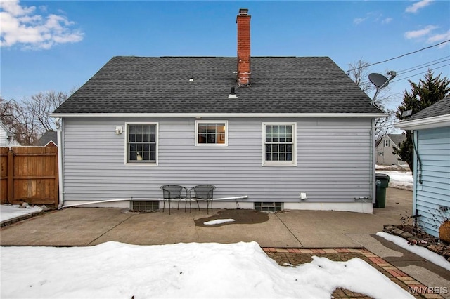 snow covered back of property with a shingled roof, a chimney, fence, and a patio