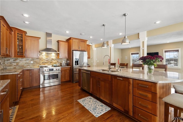 kitchen with high end appliances, glass insert cabinets, brown cabinetry, a sink, and wall chimney range hood