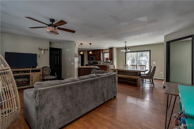 living area with dark wood-style floors, ceiling fan with notable chandelier, a baseboard heating unit, and a glass covered fireplace