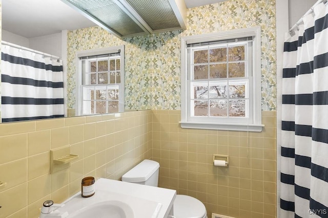 bathroom with a healthy amount of sunlight, wainscoting, toilet, and wallpapered walls