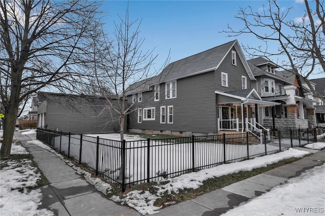 snow covered property featuring a shingled roof, a fenced front yard, and a residential view