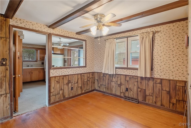 unfurnished room with wood finished floors, wainscoting, beamed ceiling, and wallpapered walls