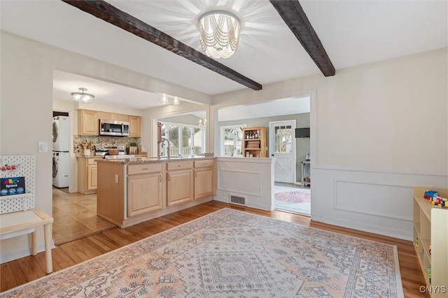 kitchen with beam ceiling, stacked washer / drying machine, stainless steel microwave, visible vents, and light brown cabinetry