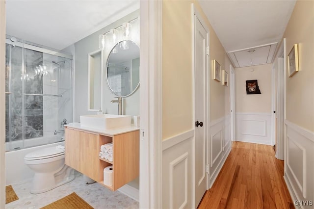 bathroom featuring toilet, a wainscoted wall, shower / bath combination with glass door, wood finished floors, and vanity