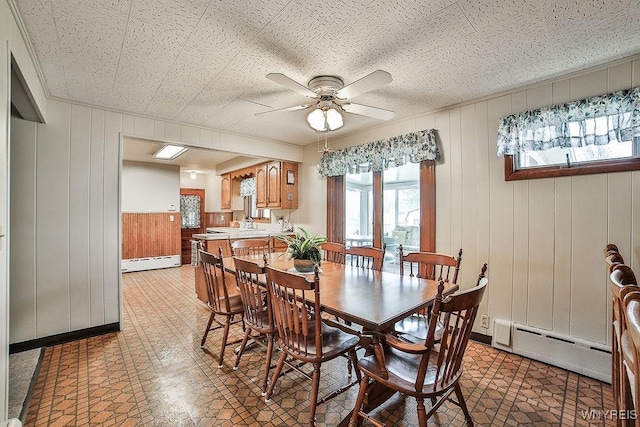 dining area with light floors, wood walls, baseboard heating, and a ceiling fan