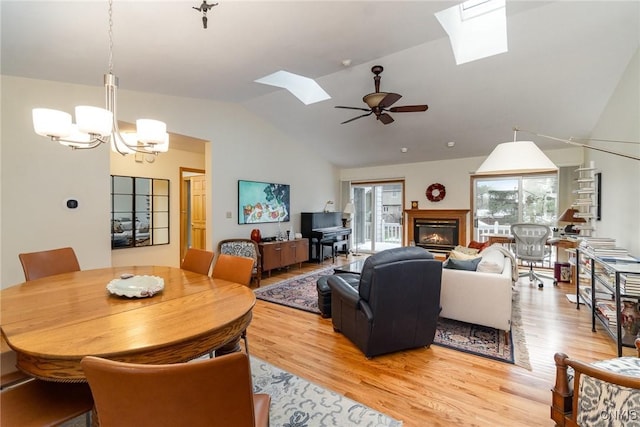 living area featuring light wood-type flooring, a glass covered fireplace, vaulted ceiling with skylight, and ceiling fan with notable chandelier