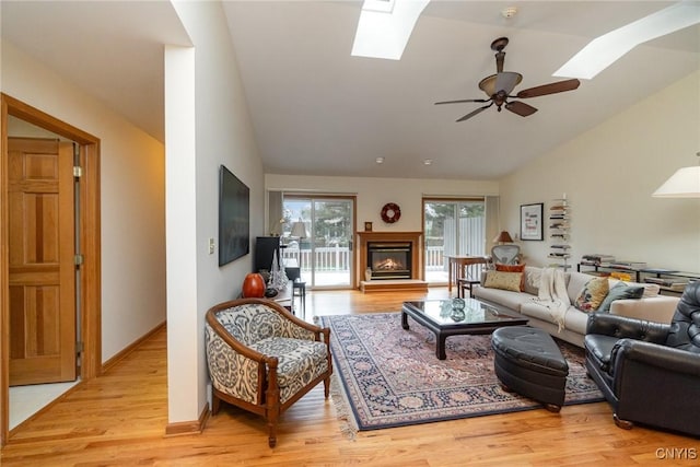 living room featuring ceiling fan, light wood-style flooring, baseboards, lofted ceiling with skylight, and a glass covered fireplace