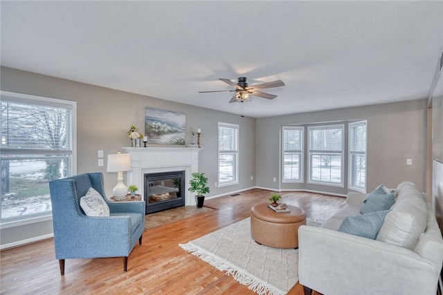 living area featuring a fireplace with flush hearth, a ceiling fan, baseboards, and wood finished floors