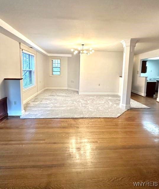 unfurnished living room featuring ornate columns, an inviting chandelier, ornamental molding, wood finished floors, and baseboards