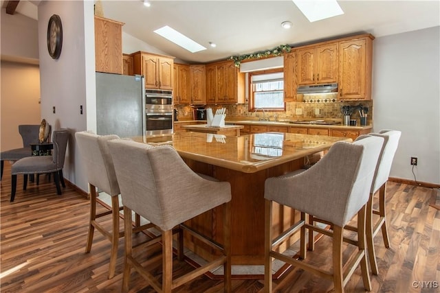 kitchen with under cabinet range hood, a kitchen island, appliances with stainless steel finishes, backsplash, and lofted ceiling with skylight