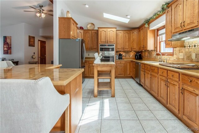 kitchen with vaulted ceiling with skylight, appliances with stainless steel finishes, under cabinet range hood, a sink, and light tile patterned flooring