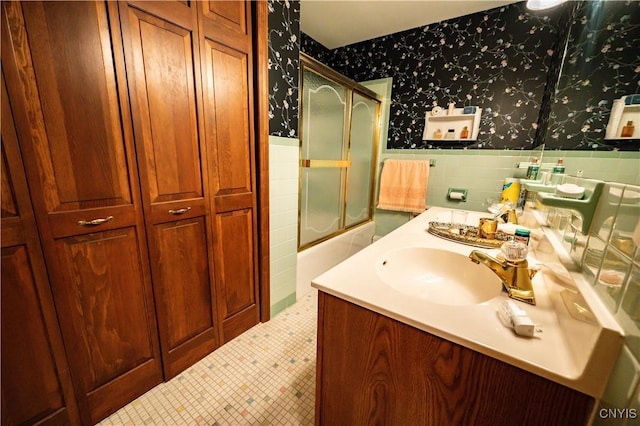 bathroom featuring wallpapered walls, shower / bath combination with glass door, a wainscoted wall, vanity, and tile walls