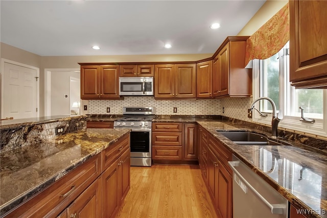 kitchen featuring brown cabinets, light wood finished floors, appliances with stainless steel finishes, a sink, and dark stone countertops