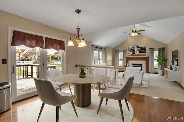 dining room featuring light wood-style floors, vaulted ceiling, a fireplace, and ceiling fan with notable chandelier