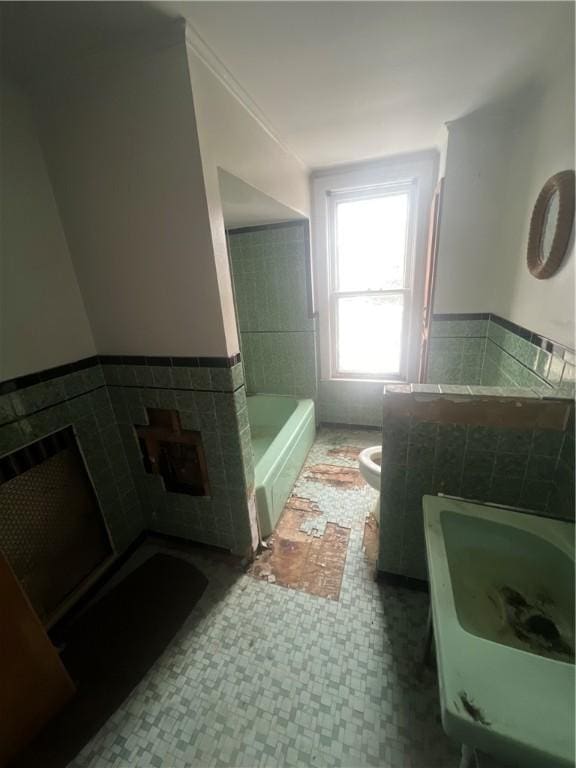 full bath featuring toilet, a bath, tile walls, and wainscoting