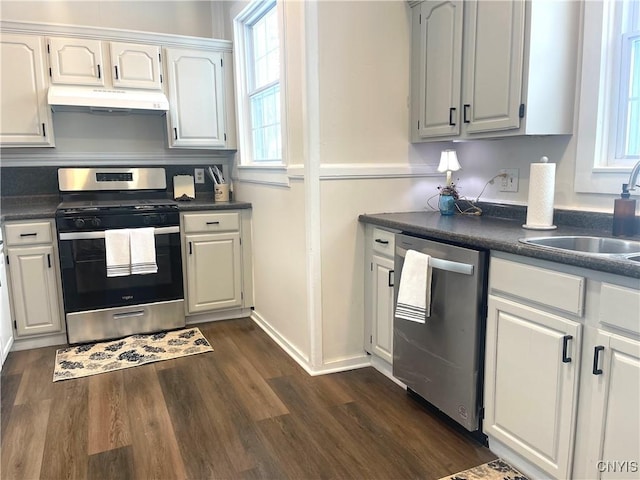 kitchen with under cabinet range hood, a sink, appliances with stainless steel finishes, dark countertops, and dark wood finished floors