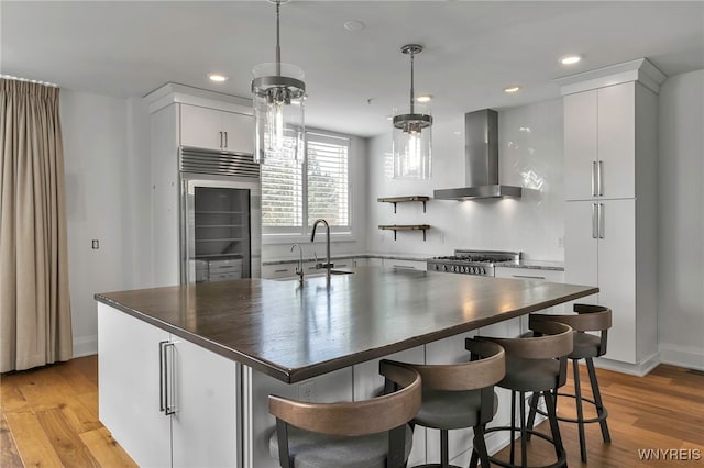 kitchen with light wood-style flooring, stove, a sink, wall chimney exhaust hood, and stainless steel built in refrigerator