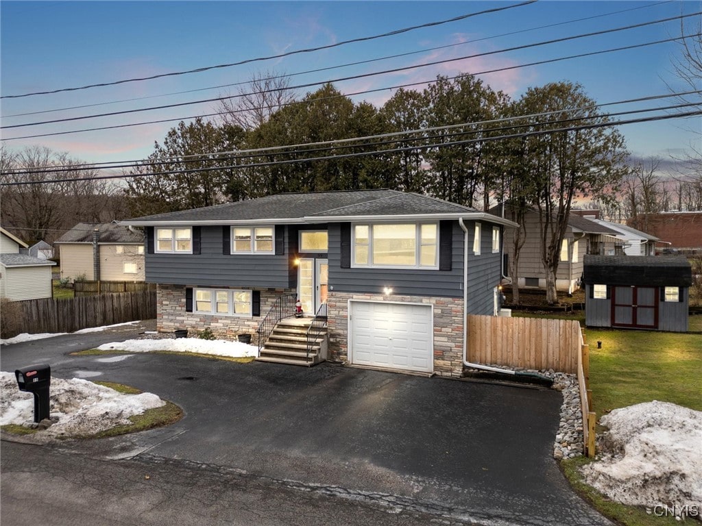 bi-level home with driveway, a garage, stone siding, fence, and a shed