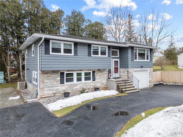 split foyer home featuring central AC unit, stone siding, aphalt driveway, an attached garage, and fence