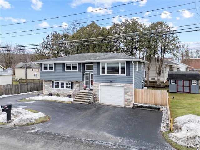 raised ranch featuring aphalt driveway, a storage shed, a garage, fence, and stone siding