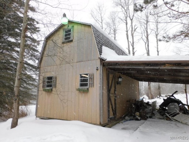 snow covered property with a carport, roof with shingles, a barn, and a gambrel roof