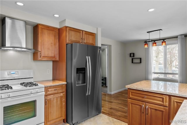 kitchen featuring light stone counters, stainless steel refrigerator with ice dispenser, wall chimney exhaust hood, brown cabinetry, and white gas range