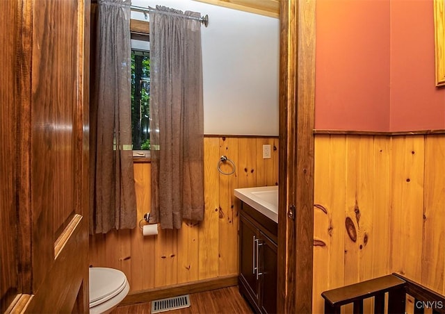 half bathroom with visible vents, toilet, wood finished floors, vanity, and wood walls