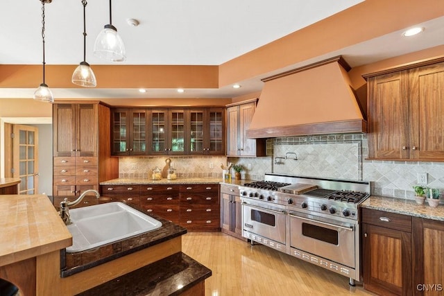 kitchen with range with two ovens, custom exhaust hood, hanging light fixtures, a sink, and light wood-type flooring