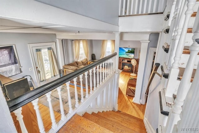 stairs with a warm lit fireplace, decorative columns, and wood finished floors