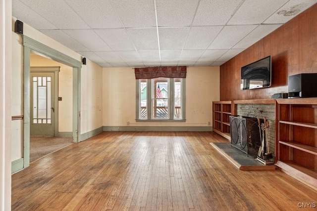 unfurnished living room featuring wood-type flooring, baseboards, a fireplace with raised hearth, and a drop ceiling