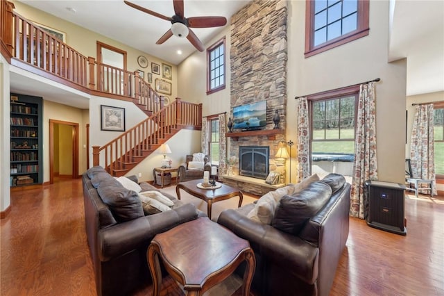 living area featuring a high ceiling, a ceiling fan, a stone fireplace, wood finished floors, and stairs