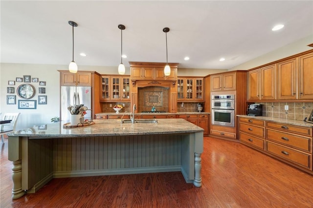 kitchen with brown cabinetry, appliances with stainless steel finishes, wood finished floors, a kitchen island with sink, and a sink