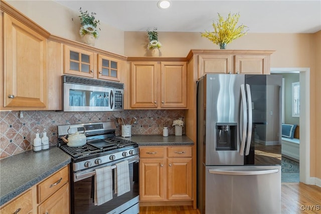 kitchen with stainless steel appliances, backsplash, light wood finished floors, dark countertops, and glass insert cabinets