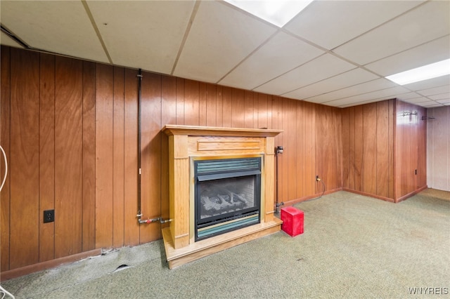 unfurnished living room featuring a paneled ceiling, carpet floors, wooden walls, and a glass covered fireplace