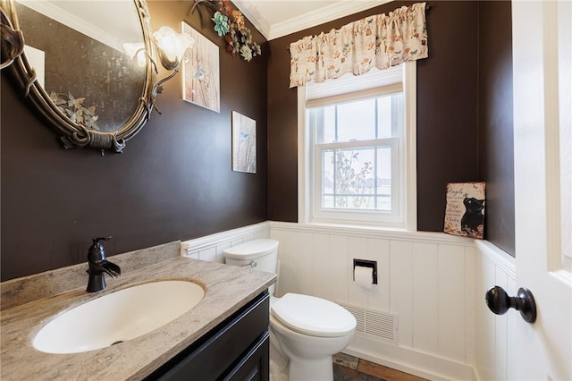 half bathroom featuring a wainscoted wall, visible vents, toilet, crown molding, and vanity