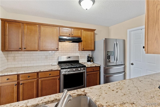 kitchen featuring light stone countertops, brown cabinetry, decorative backsplash, under cabinet range hood, and appliances with stainless steel finishes