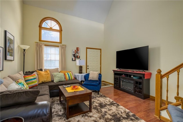 living room with high vaulted ceiling, a glass covered fireplace, wood finished floors, stairway, and baseboards