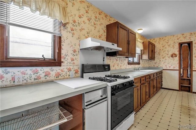 kitchen featuring under cabinet range hood, light floors, wallpapered walls, white range with gas stovetop, and a healthy amount of sunlight