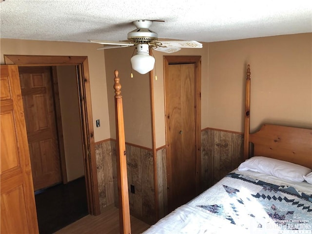 bedroom with a textured ceiling, wooden walls, a ceiling fan, and wainscoting