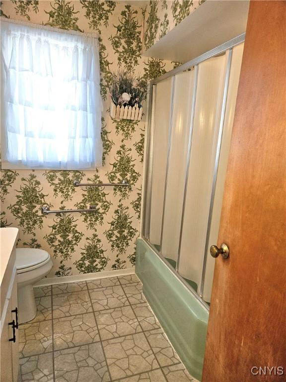 bathroom featuring toilet, shower / bath combination with glass door, wallpapered walls, baseboards, and vanity