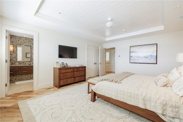 bedroom featuring a raised ceiling, baseboards, light wood finished floors, and connected bathroom