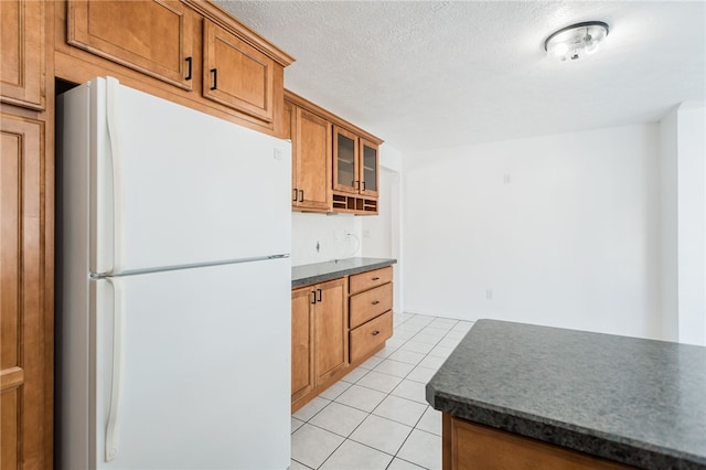 kitchen with dark countertops, glass insert cabinets, light tile patterned floors, freestanding refrigerator, and brown cabinetry