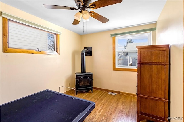 exercise area featuring visible vents, baseboards, a wood stove, wood finished floors, and a ceiling fan