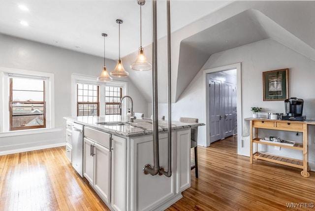 kitchen with light wood finished floors, a kitchen island with sink, light stone countertops, stainless steel dishwasher, and a sink