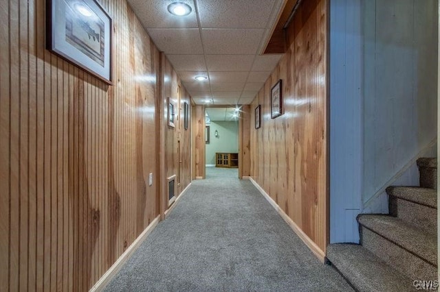 hall with stairway, wooden walls, baseboards, a drop ceiling, and carpet flooring