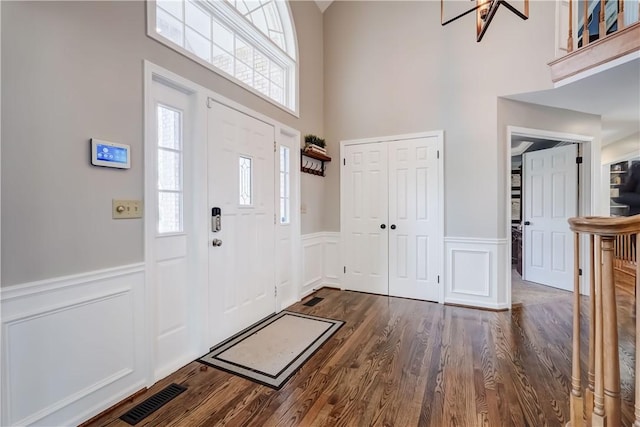 entrance foyer with a wainscoted wall, dark wood-type flooring, visible vents, and a towering ceiling